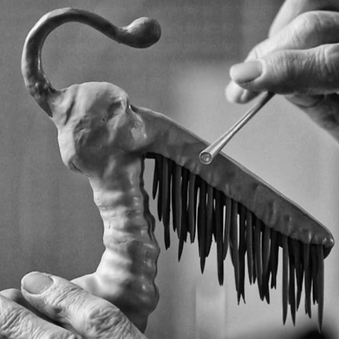H&auml;nde halten eine skulpturartige Figur mit einem gebogenen Kopf und einem kammartigen K&ouml;rper, w&auml;hrend ein St&auml;bchen darauf zeigt.